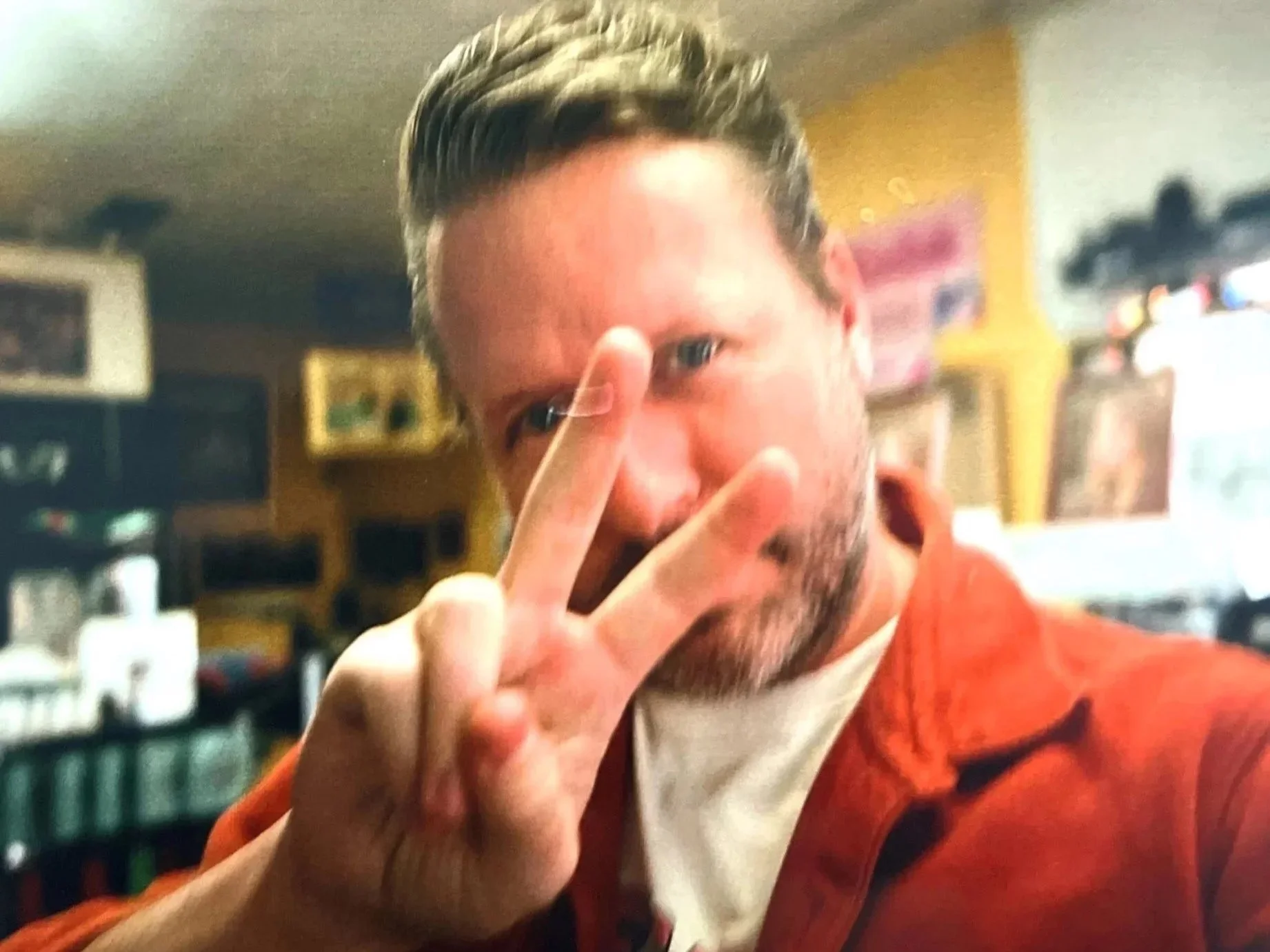 A man with short, styled hair making a peace sign near his face while looking at the camera, in a cozy indoor setting with shelves and electronic devices in the background.