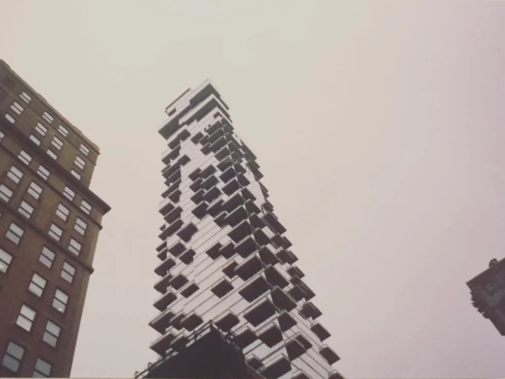 A tall, modern skyscraper with a unique, stacked design in an urban setting, surrounded by other buildings under a cloudy sky.