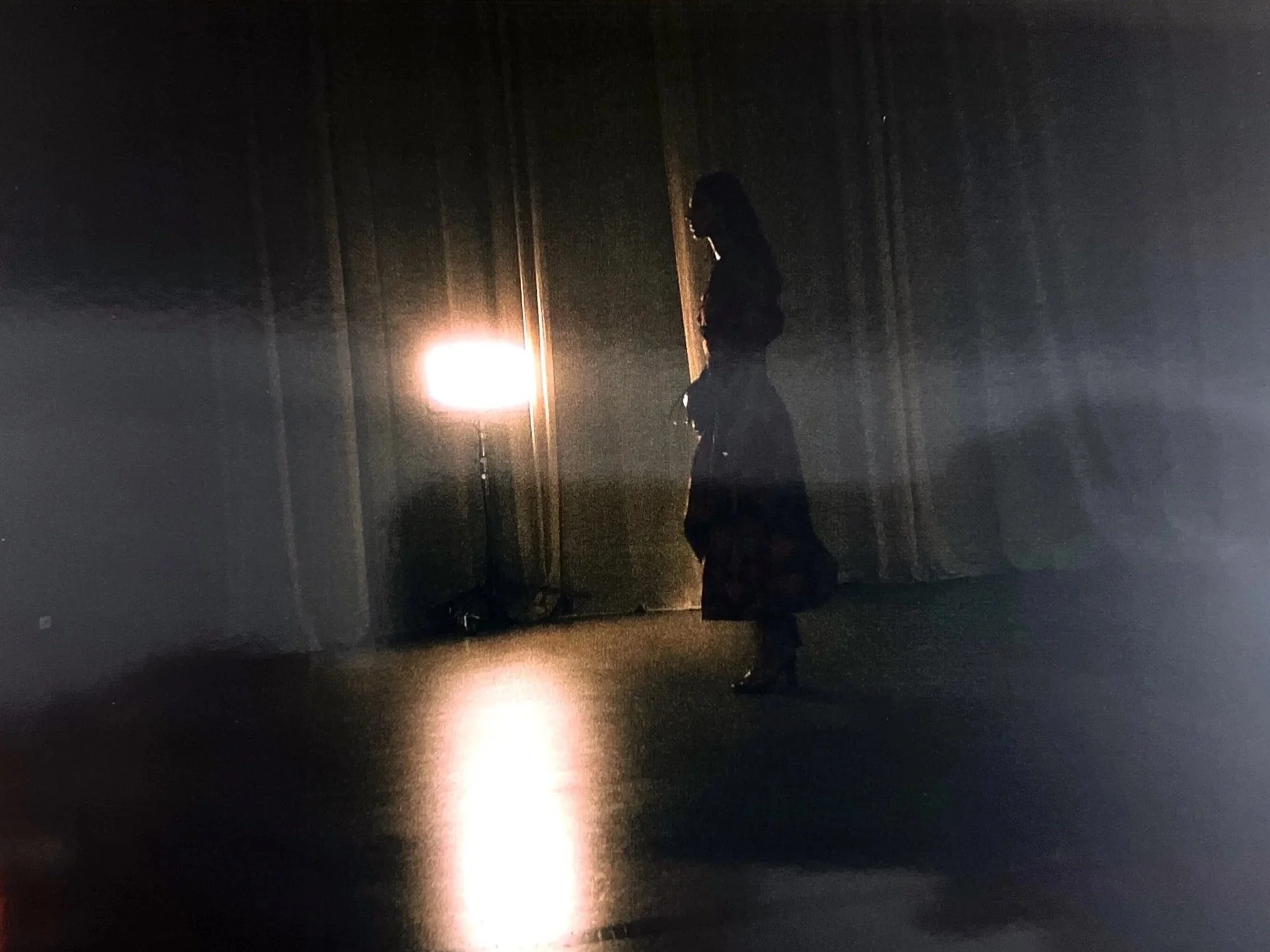 A person in silhouette standing on a dark stage with a bright spotlight behind them, creating a glow and lens flare.