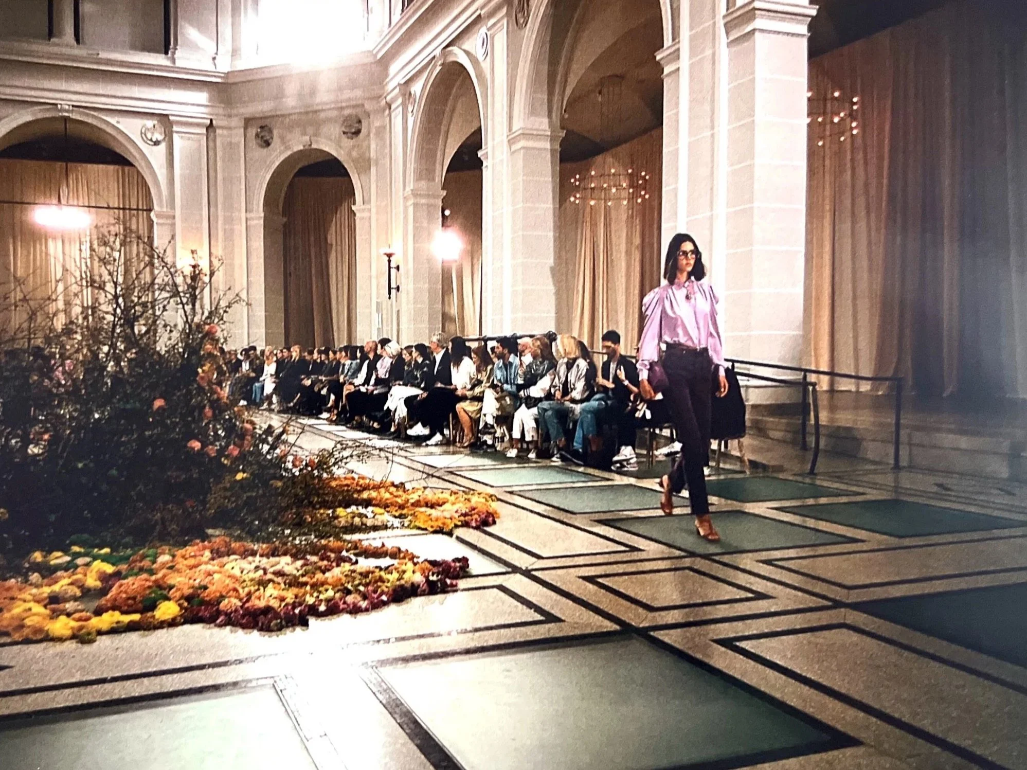 A woman in a pink blouse and black pants walking past a seated audience in a grand hall with high arching columns and draped curtains. There is a floral arrangement in the foreground.
