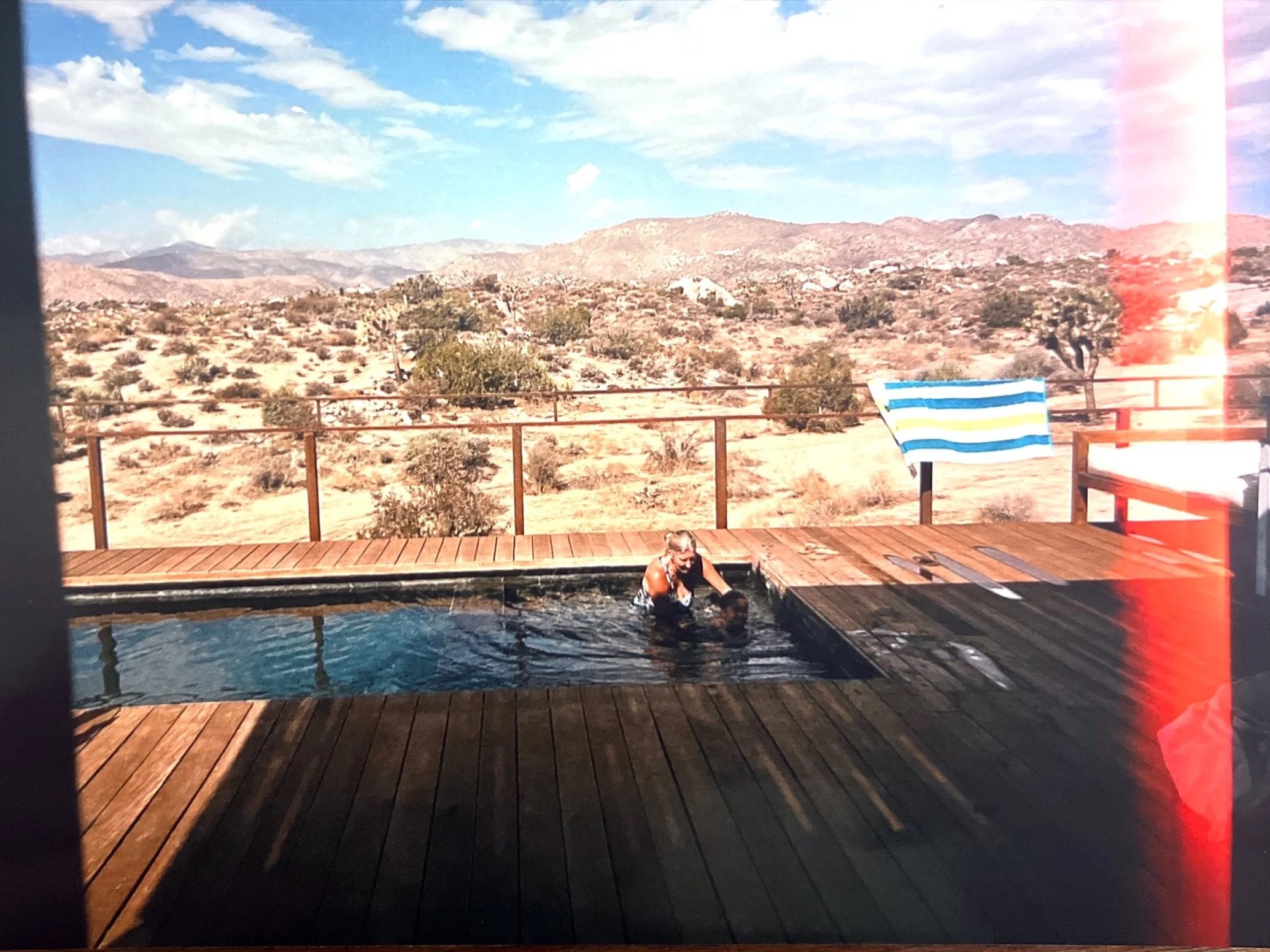 Person swimming in an outdoor pool with a desert landscape and mountains in the background, blue sky with scattered clouds, and a striped towel on a lounge chair.