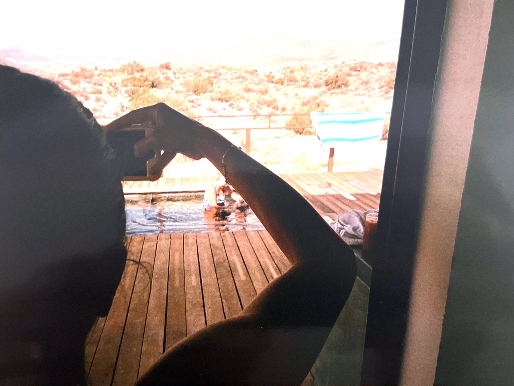 Person taking a selfie on a wooden deck with a hot tub, a person floating in the hot tub, and a view of a natural landscape with trees and hills in the background.