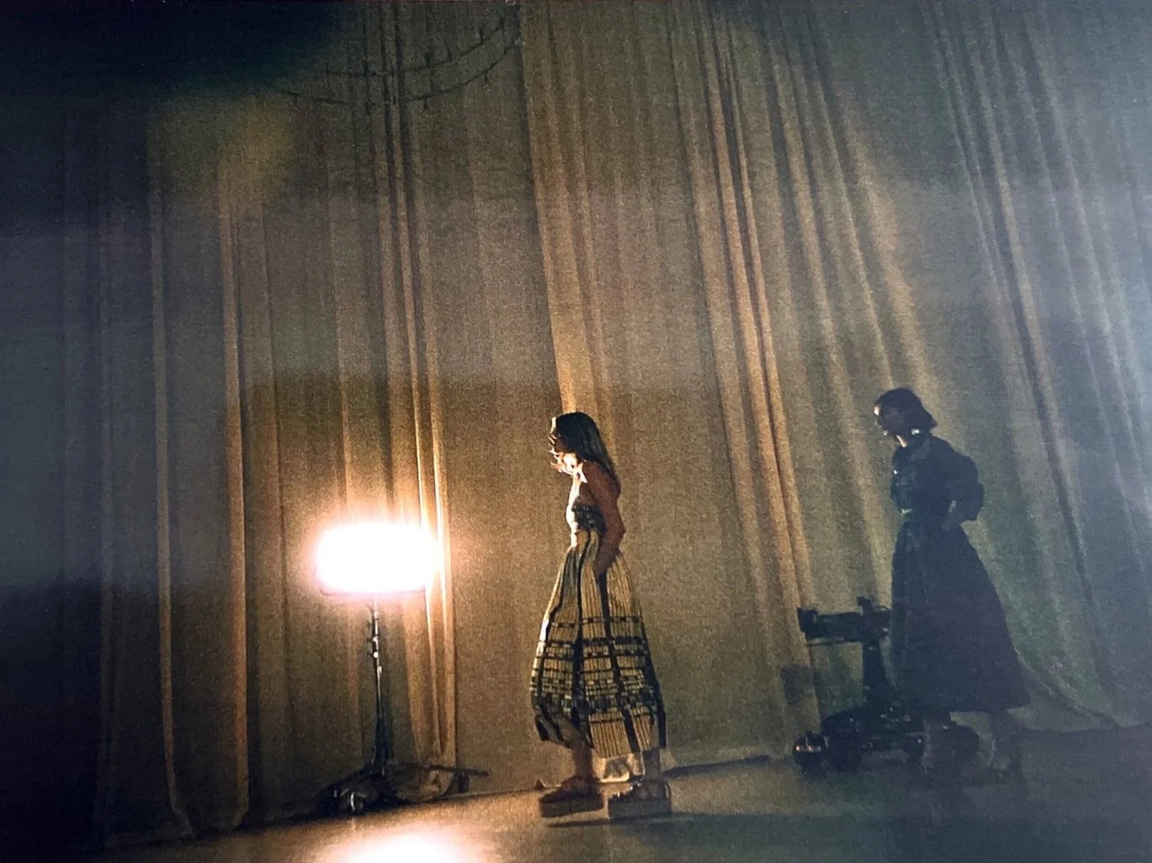 A woman in a long skirt and a mask stands next to a bright lamp on a stand, with another woman in dark clothing and glasses standing nearby, both against a large curtain backdrop.