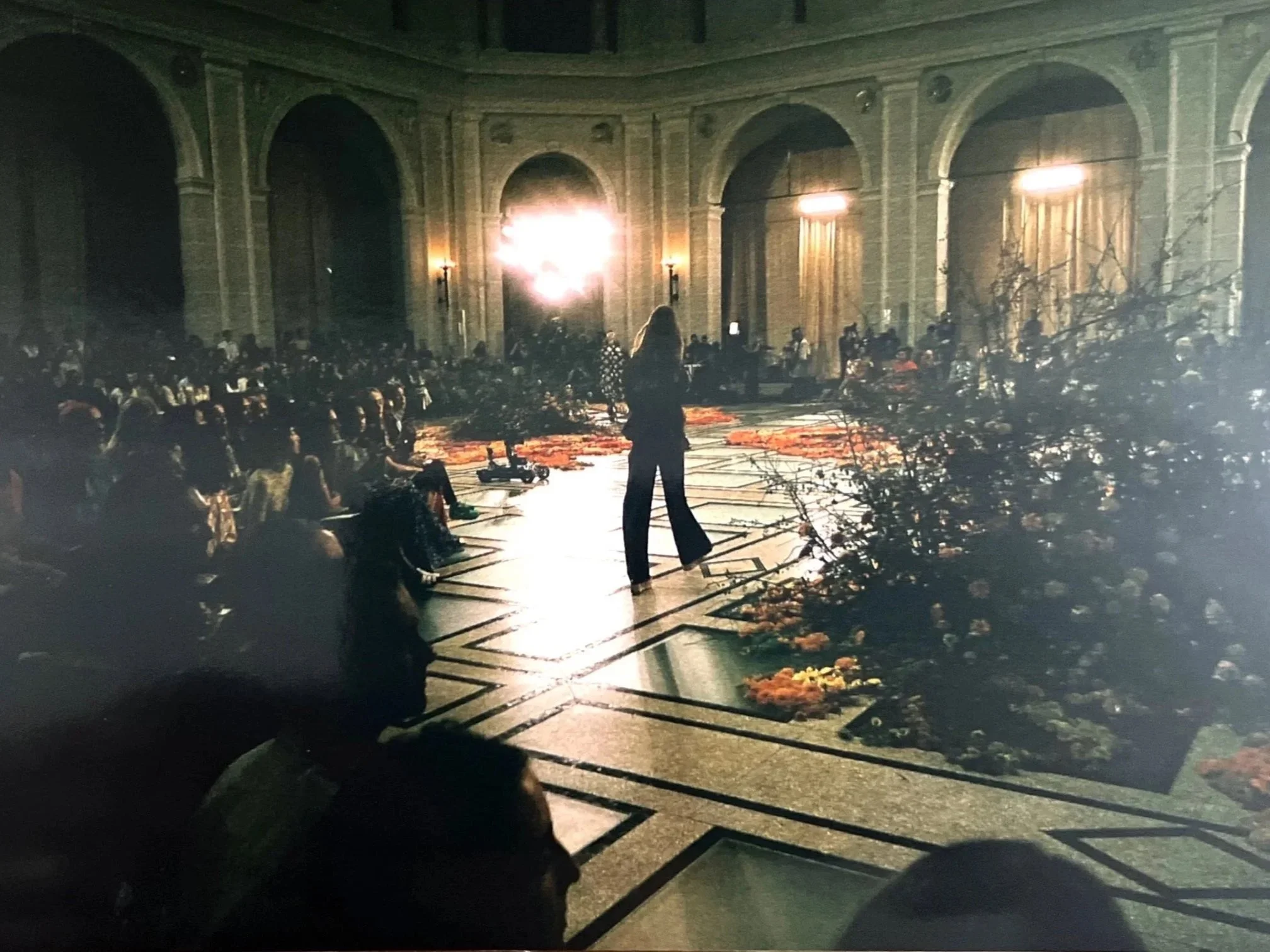 A woman stands on a stage in front of a seated audience in a large, ornate hall with high arches. The scene is illuminated by bright lights.