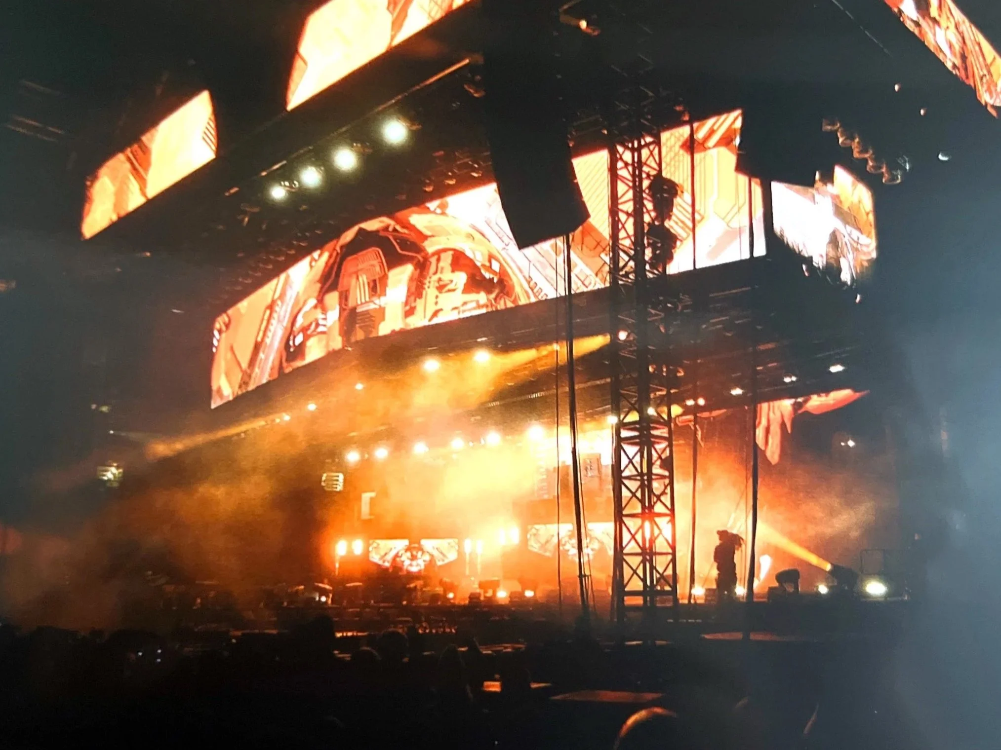 A concert stage with large screens displaying orange graphics, bright orange stage lights, and a few silhouette figures including a person with a camera.