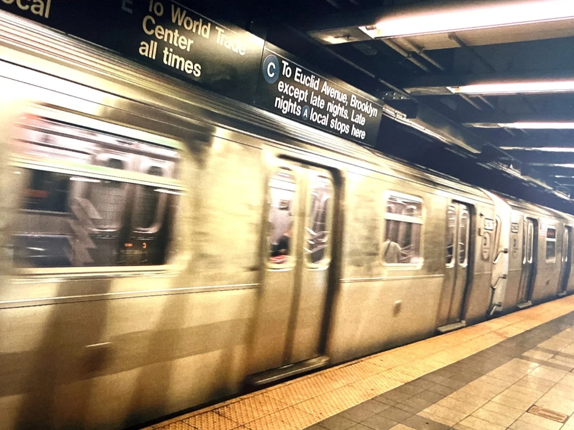 A subway train arriving at a station platform with an overhead sign showing directions to Brooklyn and Euclid Avenue. The platform has a yellow safety strip, and the train has a metallic exterior with windows reflecting the surroundings.