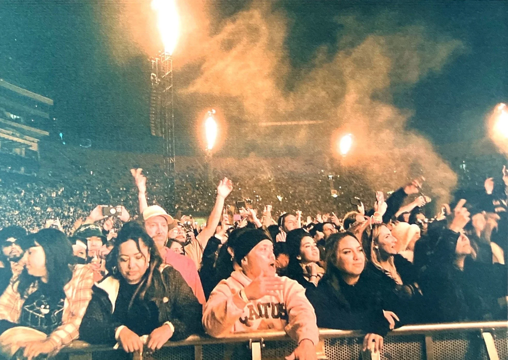 Crowd of people at a concert or event, some taking photos or videos, with fireworks lighting up the sky in the background.