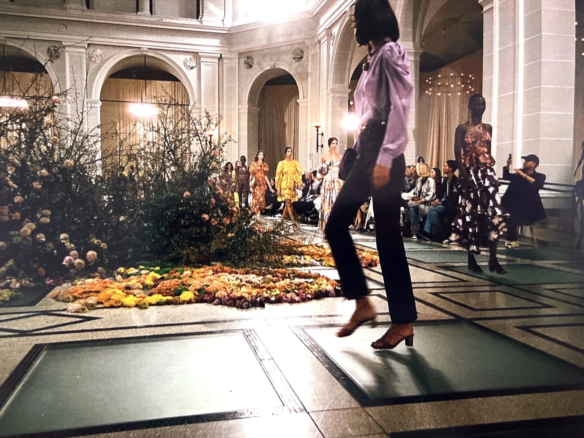Fashion models walk on a runway with floral decorations in an elegant grand hall, with an audience seated on either side.