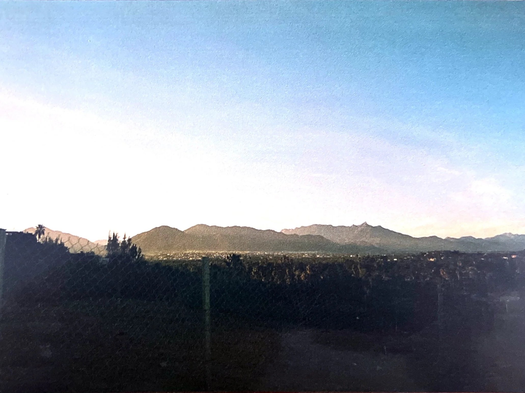 View of distant mountain range against a twilight sky, with a dark silhouette of trees and a fence in the foreground.