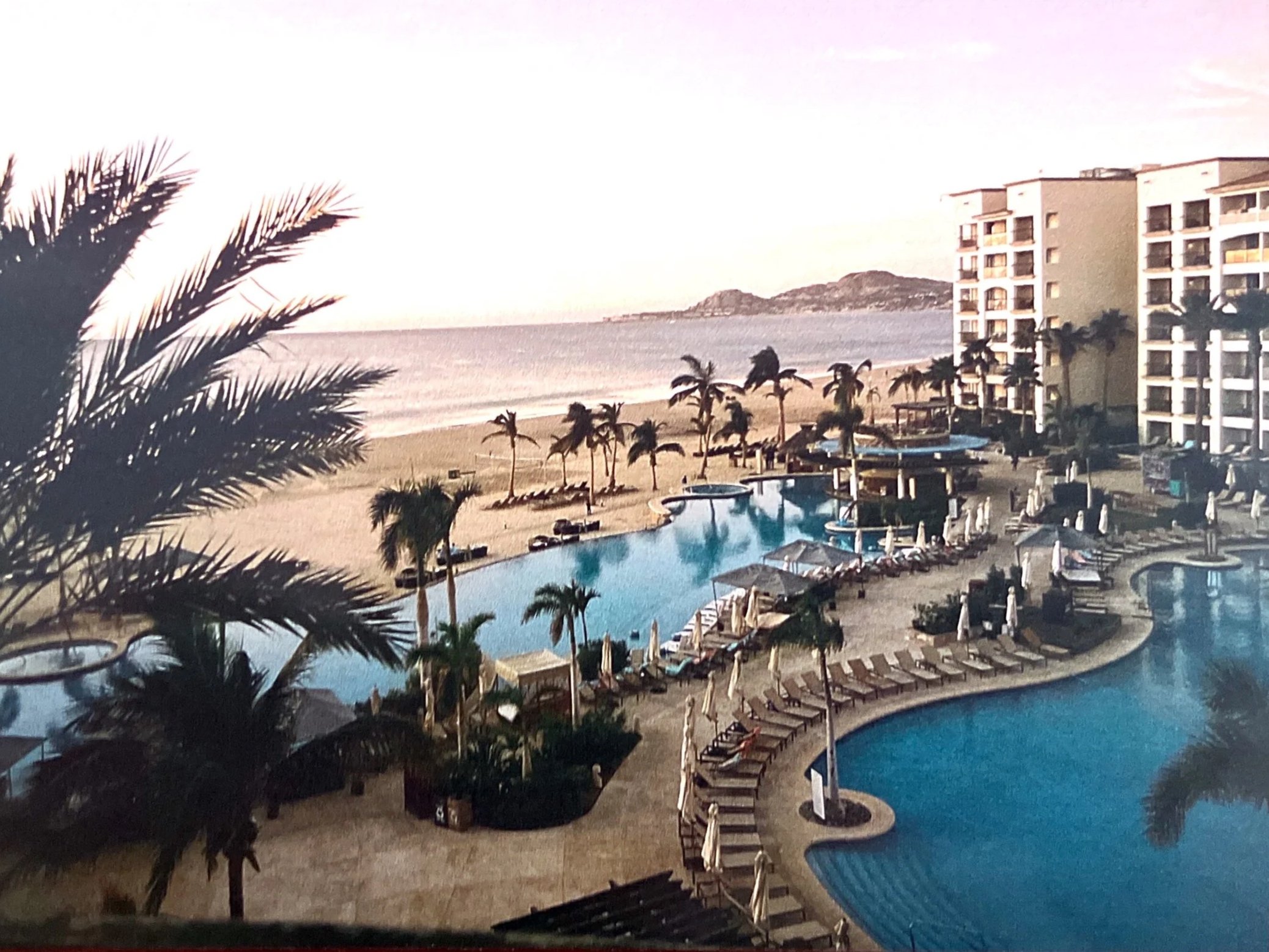 View of a beachside resort with a large swimming pool, surrounded by lounge chairs and umbrellas, overlooking the ocean with palm trees and a distant hill.