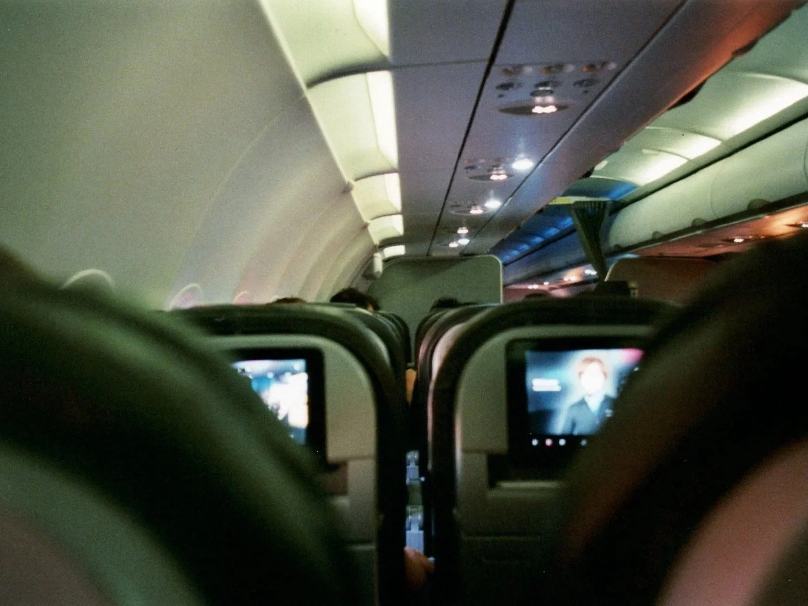 Inside an airplane cabin viewed from the back seats showing overhead compartments, some passengers and in-flight entertainment screens.