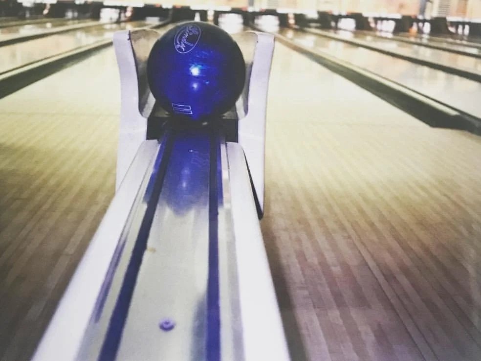 A blue bowling ball on a metal ball return track at a bowling alley, with several bowling lanes in the background.