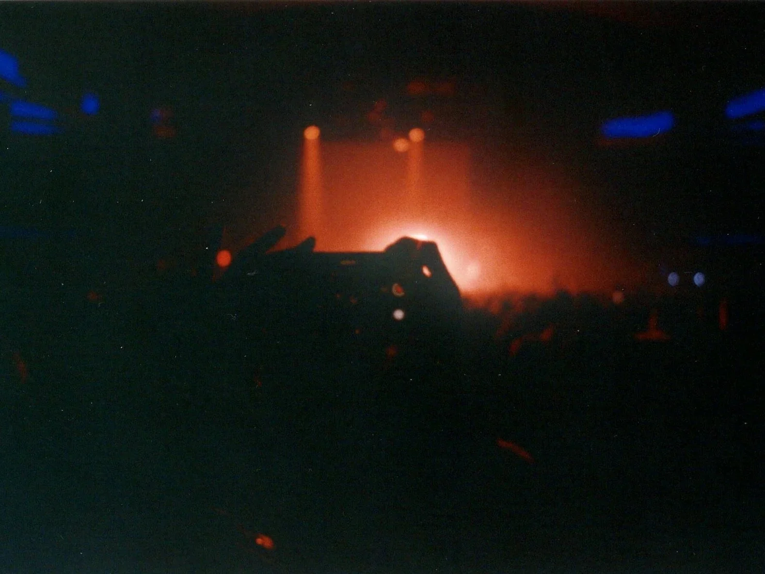 Silhouette of a person's hand making a peace sign in front of colorful stage lights at a concert or nightclub.