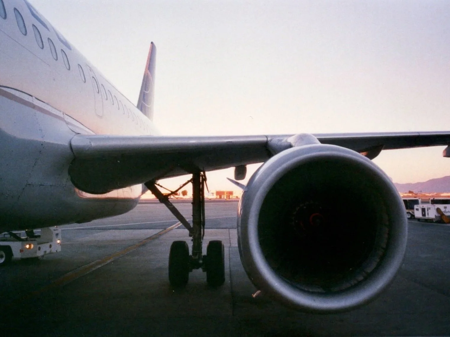 Close-up of an airplane's engine and wing on the tarmac with other airport vehicles in the background during sunset.