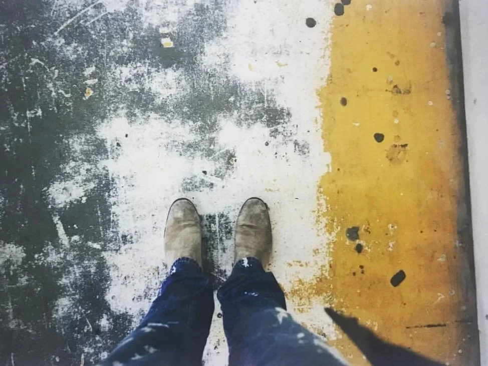 Top-down view of a person wearing tan work boots and blue jeans standing on a worn concrete floor with black and yellow markings.