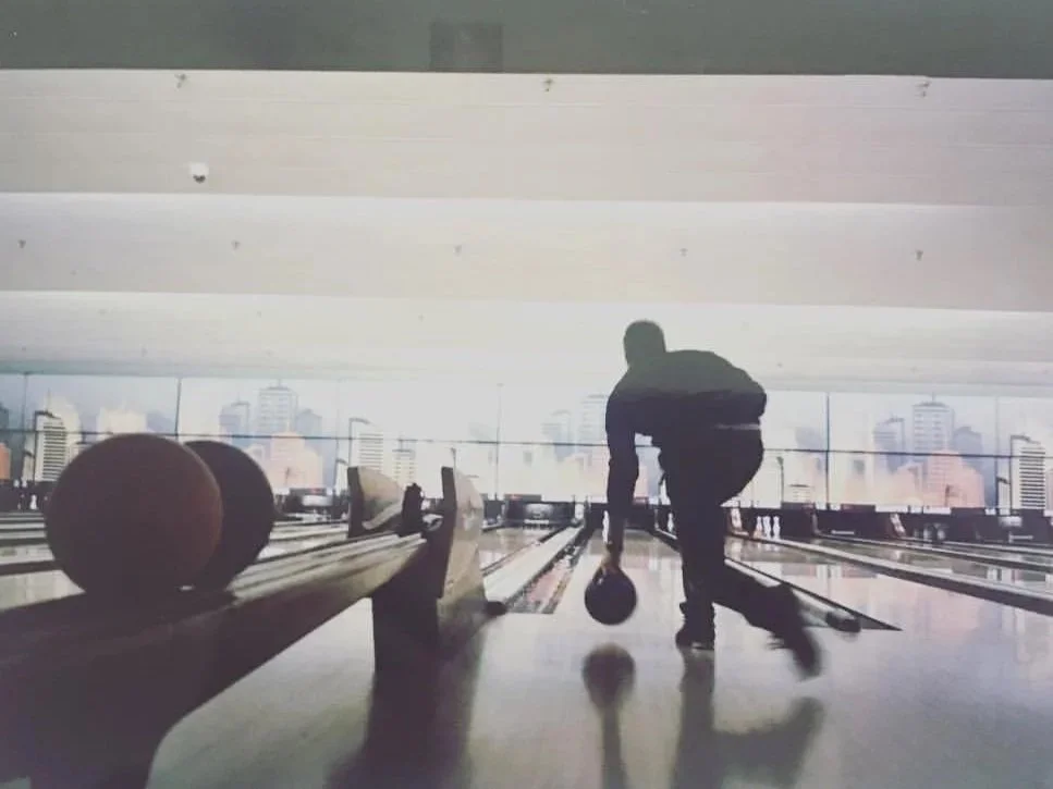 A man at a bowling alley is picking up a bowling ball, with several other balls on the ball return. The alley has a view of a city skyline through large windows.