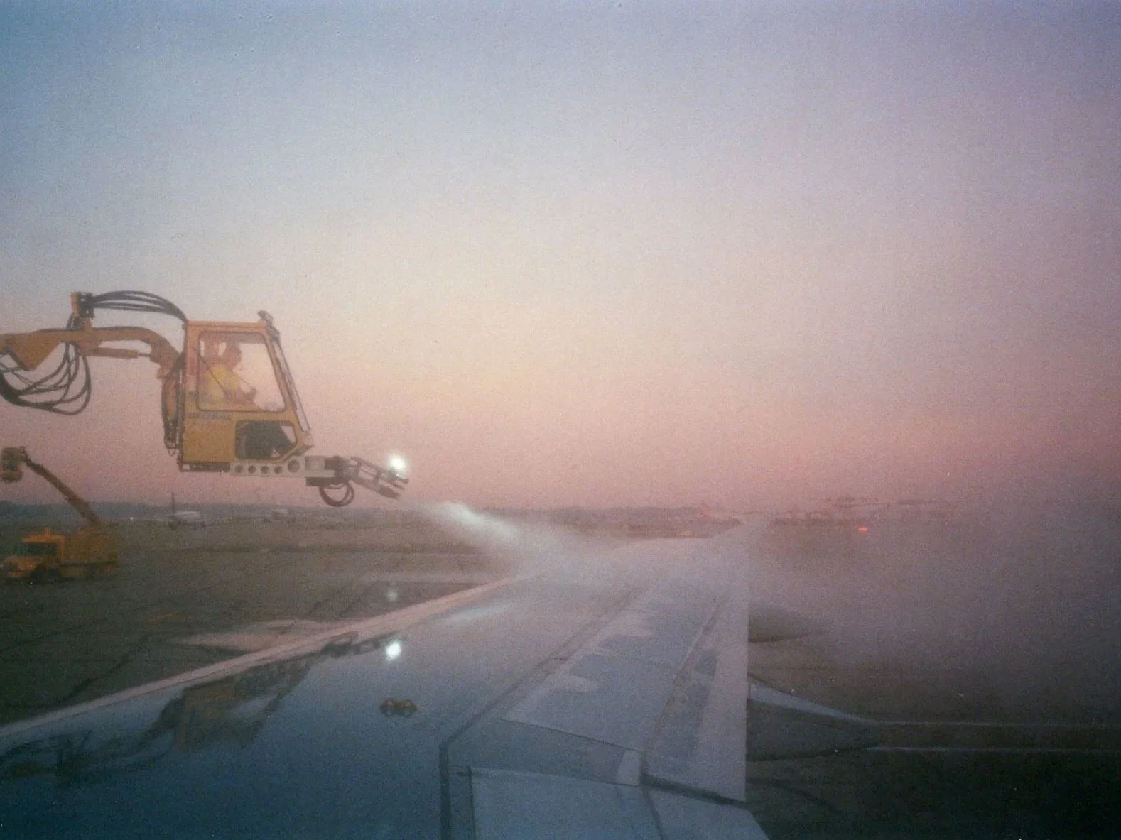 View from an airplane window showing a construction vehicle on the runway during a foggy or dusty evening or early morning.