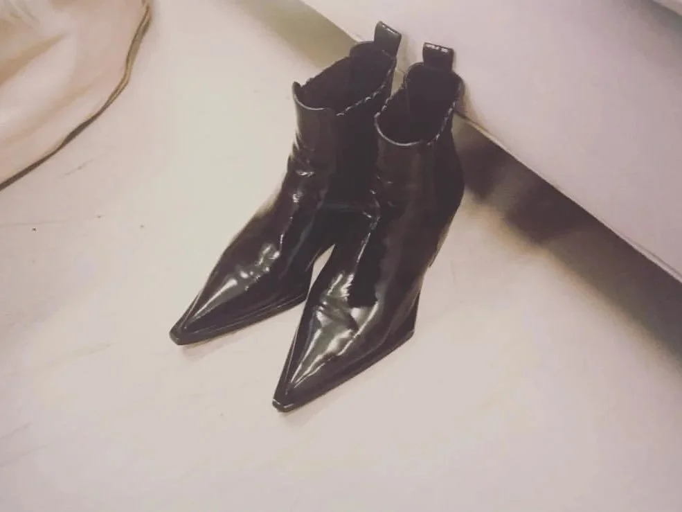 A pair of shiny black high-heeled ankle boots with pointed toes placed on a light-colored floor near a piece of furniture.