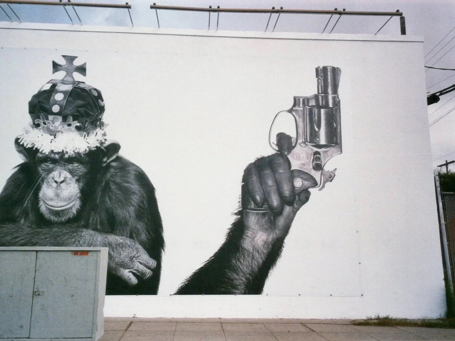A black and white mural of a chimpanzee wearing a crown with a cross on top, holding a revolver in its right hand, painted on a white wall.