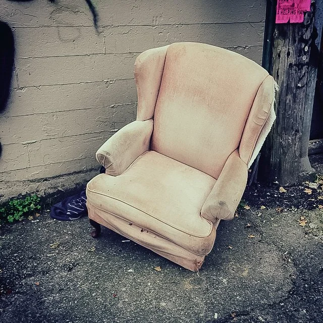 An old beige armchair with worn upholstery positioned outdoors next to a wooden utility pole and a brick wall.