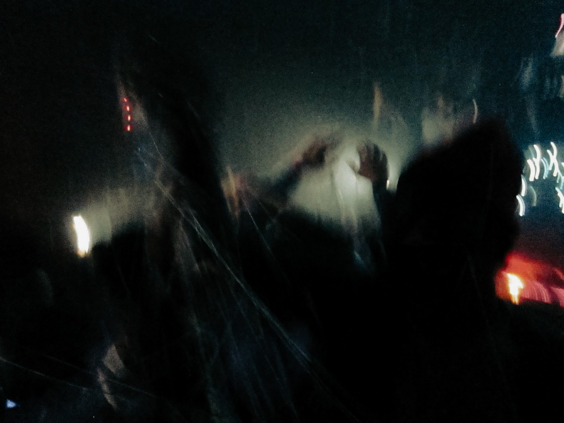 The image shows a blurry nighttime scene through a vehicle's windshield, with raindrops and streaks of rain visible. The interior silhouette of a person is also visible, likely the driver.