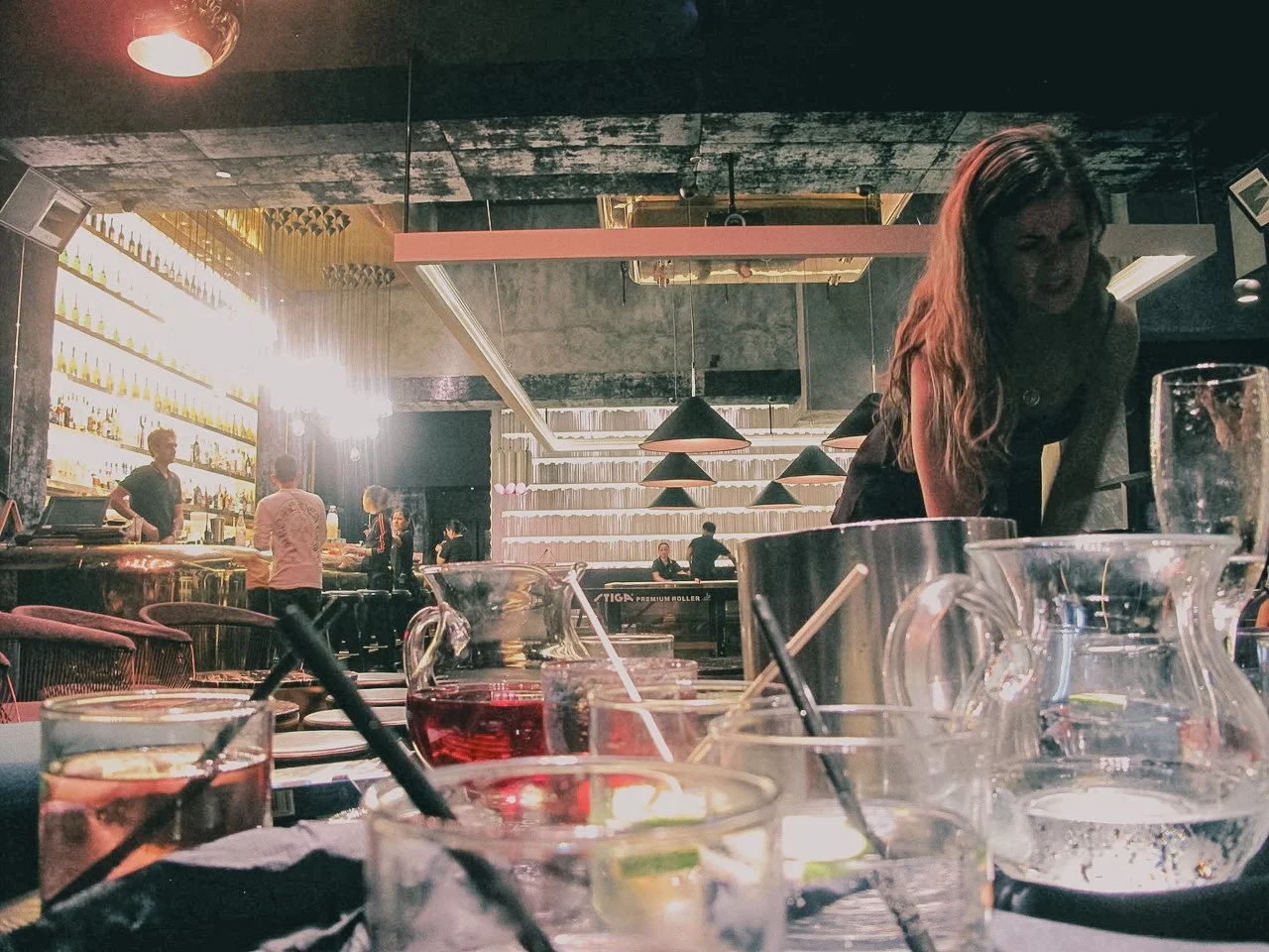 Interior of a modern bar or restaurant with drinks on the table, a woman leaning over a table, and people in the background near a bar counter and shelving with bottles.