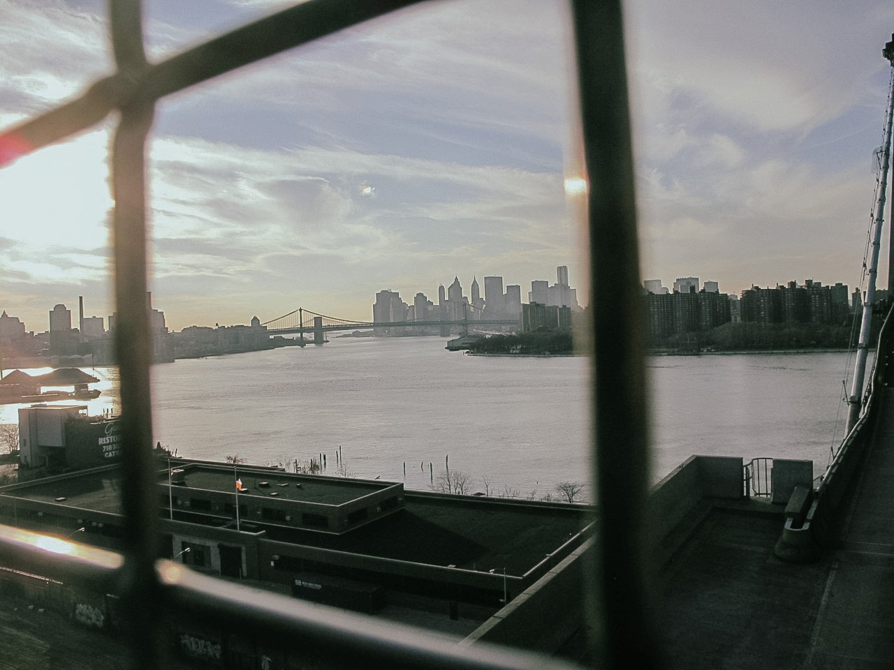 View of the Manhattan skyline with skyscrapers and the Brooklyn Bridge across the East River, seen through a window with metal grid bars.