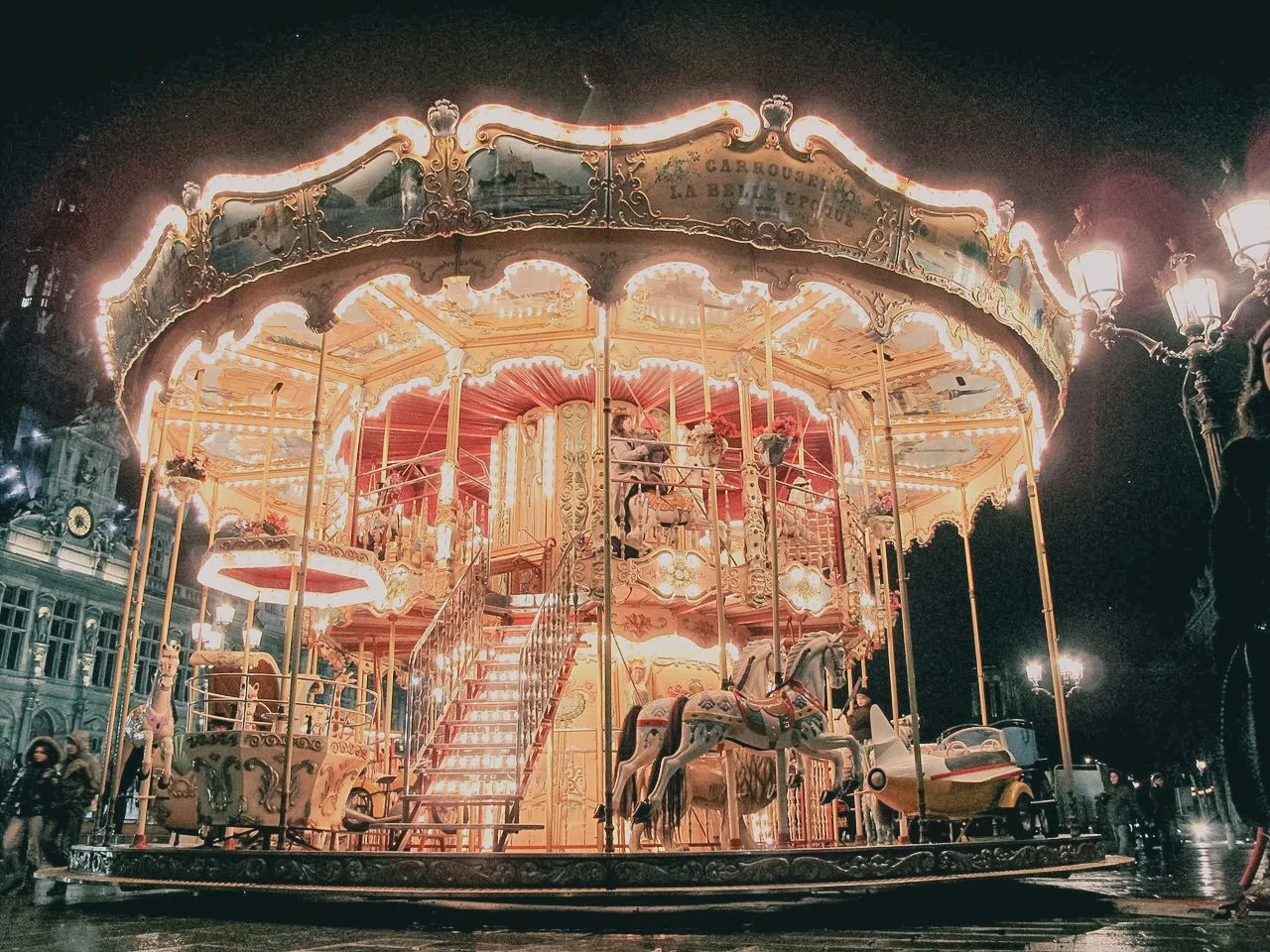 Illuminated vintage carousel with ornate details and horses at night in a city square.