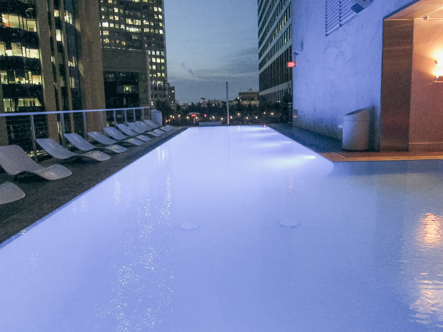 A rooftop swimming pool during dusk, with lounge chairs along one side and city buildings in the background.