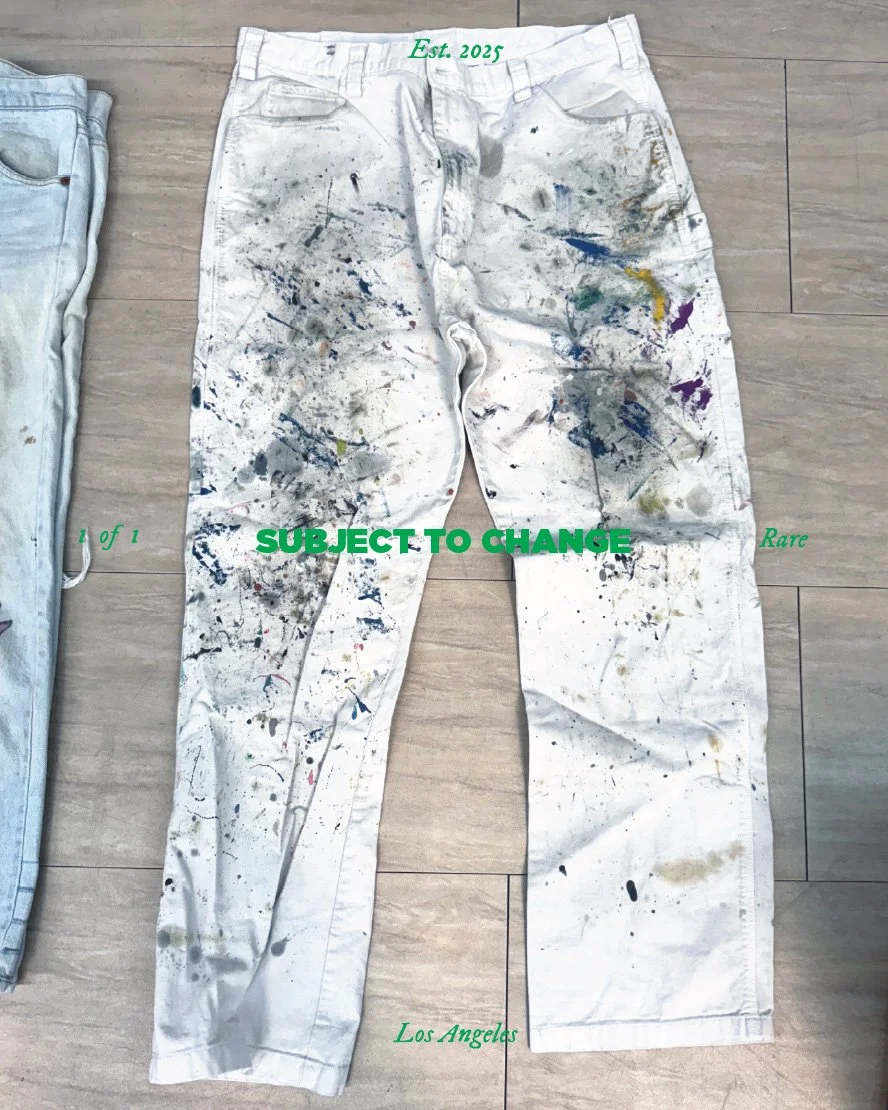 White paint-splattered jeans laying flat on a wooden floor, with green text overlay reading 'Est. 2025' at the waist and 'SUBJECT TO CHANGE' across the thighs, and smaller green text reading '1 of 1' on the left hip, 'Rare' on the right hip, and 'Los