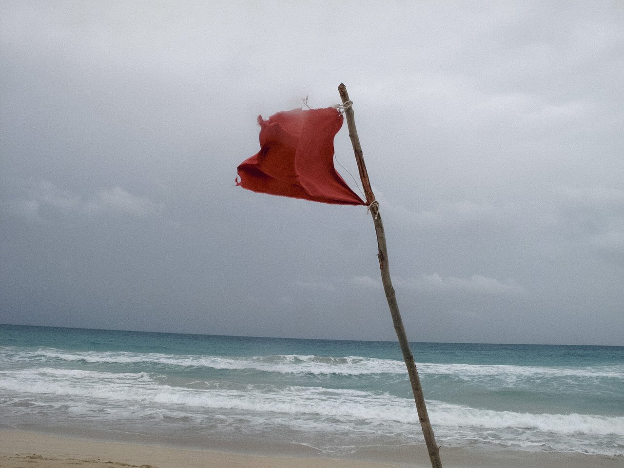 A worn red flag attached to a wooden stick on a sandy beach with ocean waves and overcast sky in the background.