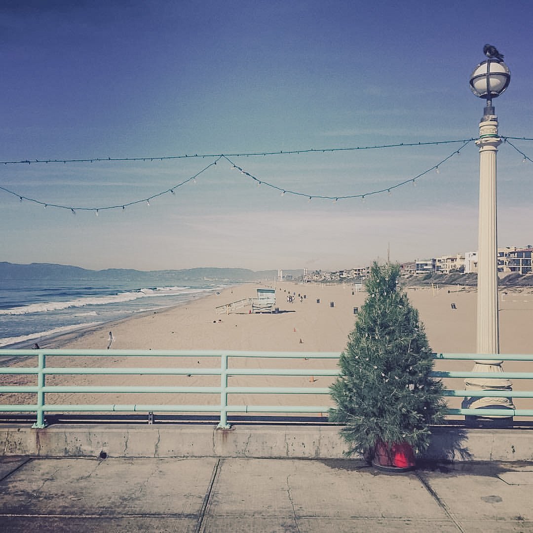A beach scene with a sidewalk in the foreground featuring a small decorated Christmas tree in a red pot, a street lamp, and string lights overhead. The sandy beach extends to the ocean with gentle waves, and buildings are visible in the distance unde