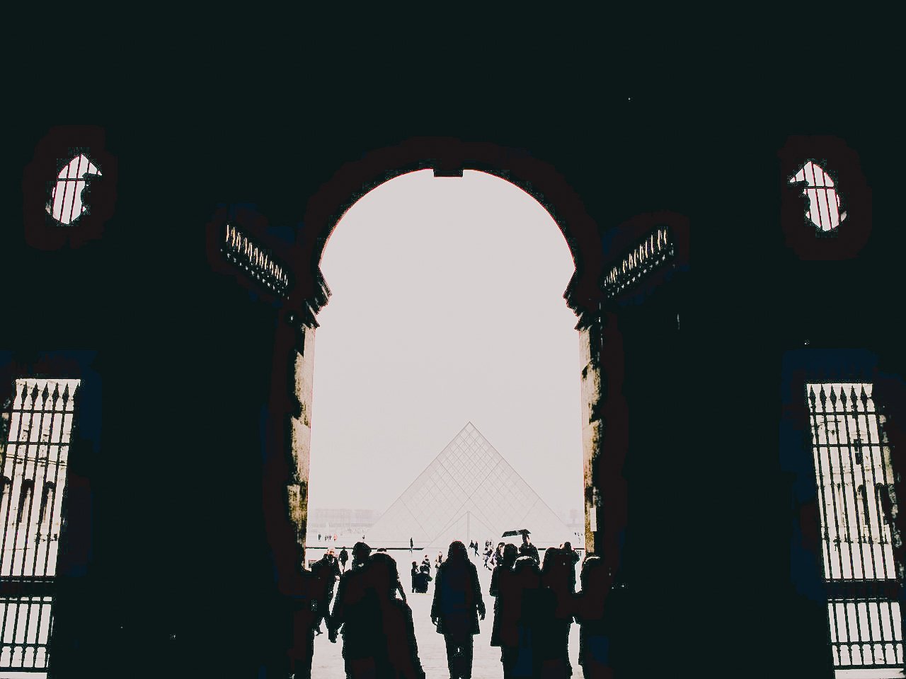 View through a large arched doorway showing the silhouette of visitors walking out to outdoor space with a glass pyramid structure in the distance.