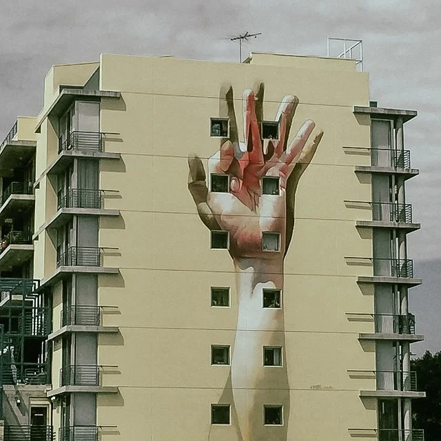 A large mural of a human hand making the OK gesture is painted on the side of a multi-story beige apartment building with small balconies on each side. The mural is designed to fit within the building's windows, creating an illusion of the hand reach
