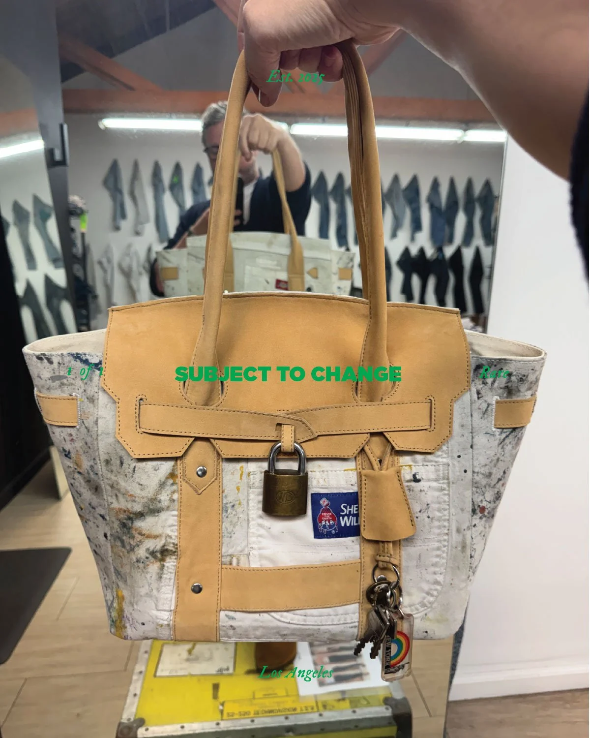 A tan and white tote bag with paint splatter design, hanging from a person's hand, in front of a mirror in a knife shop, with a reflection of the person taking a photo. Text overlay says 'Est. 2025' at the top, 'SUBJECT TO CHANGE' in green across the