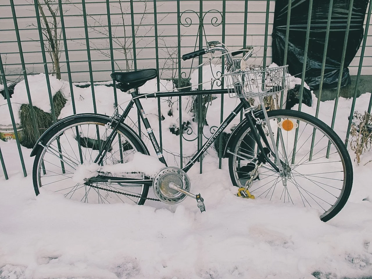 A black bicycle with a front basket is locked to a fence in a snowy outdoor area.