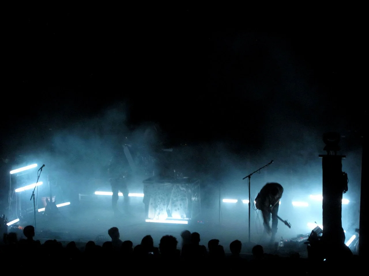 A dark stage with blue lighting, a group of musicians performing with one playing the guitar, and an audience in the foreground.
