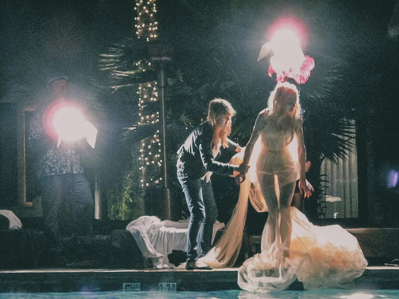 Performers on stage under bright lights, one woman in a wedding dress and others helping her, surrounded by palm trees and decorative lighting.