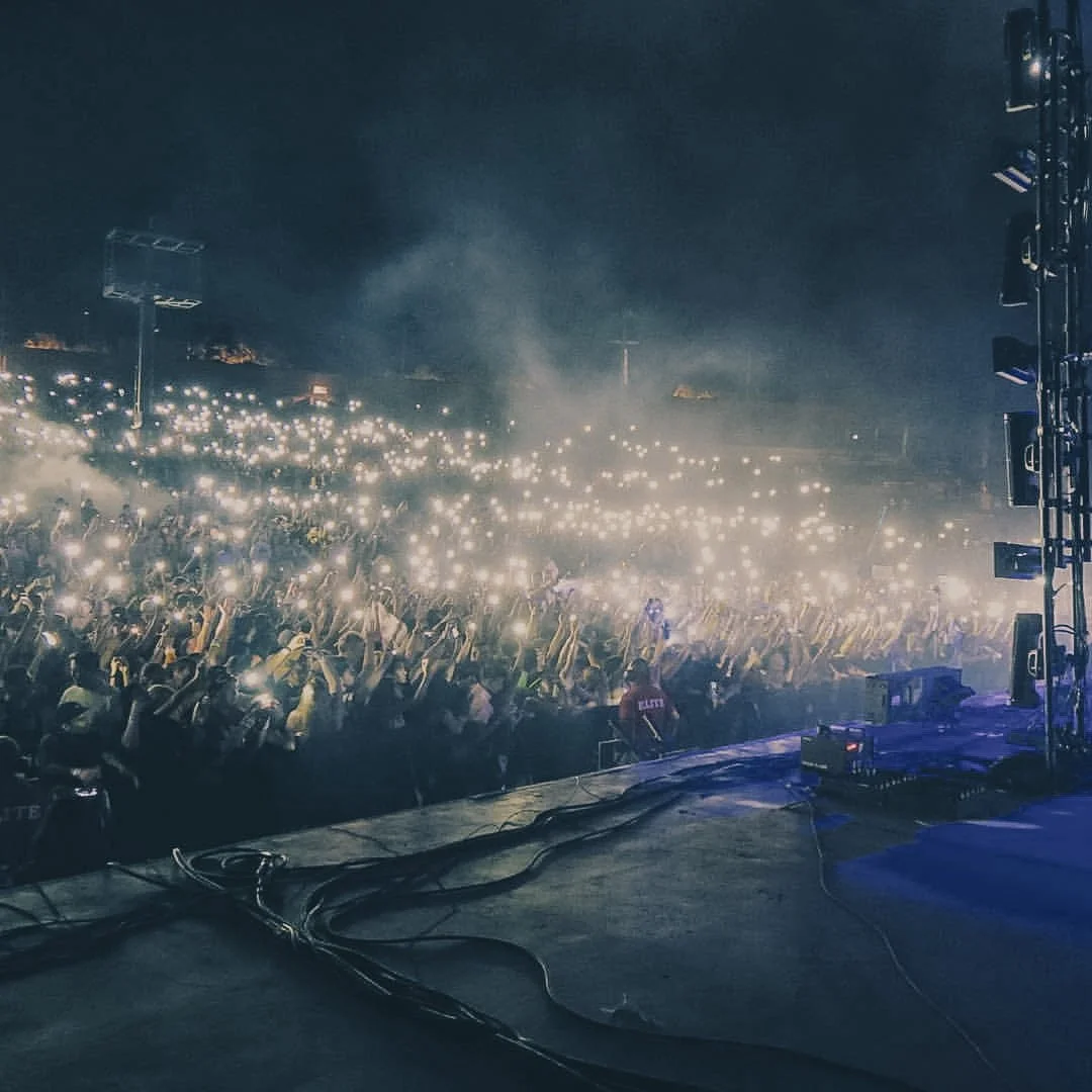 Crowd at a concert or music event with many people holding up phones with flashlight on, creating a starry light effect, seen from the stage.