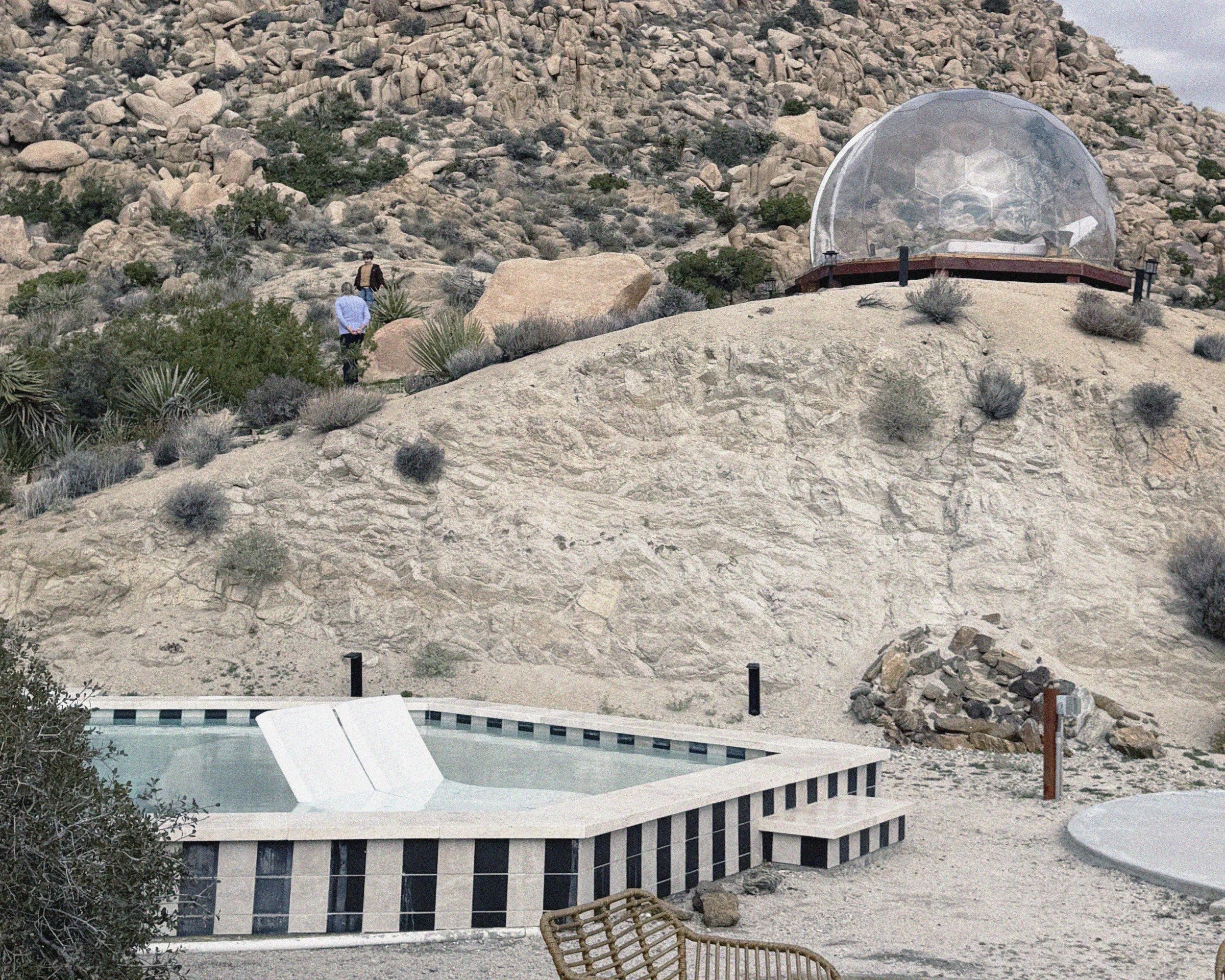 Desert landscape with rocky terrain, sparse shrubs, with a small hot tub and a clear geodesic dome on a hill, and three people walking in the background.