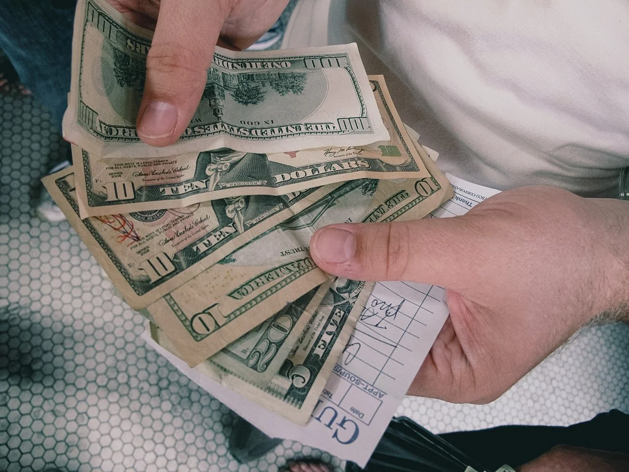 Person counting or handling multiple U.S. dollar bills, including a ten-dollar bill and others, on a clipboard with a paper receipt or form.
