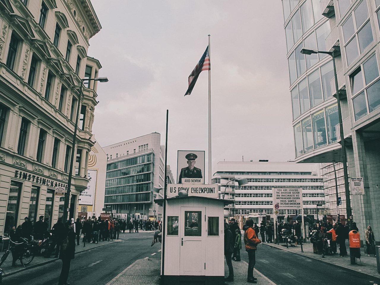 U.S. Army checkpoint booth with a sign and uniformed guard, surrounded by people on a city street with modern buildings and flags in the background.