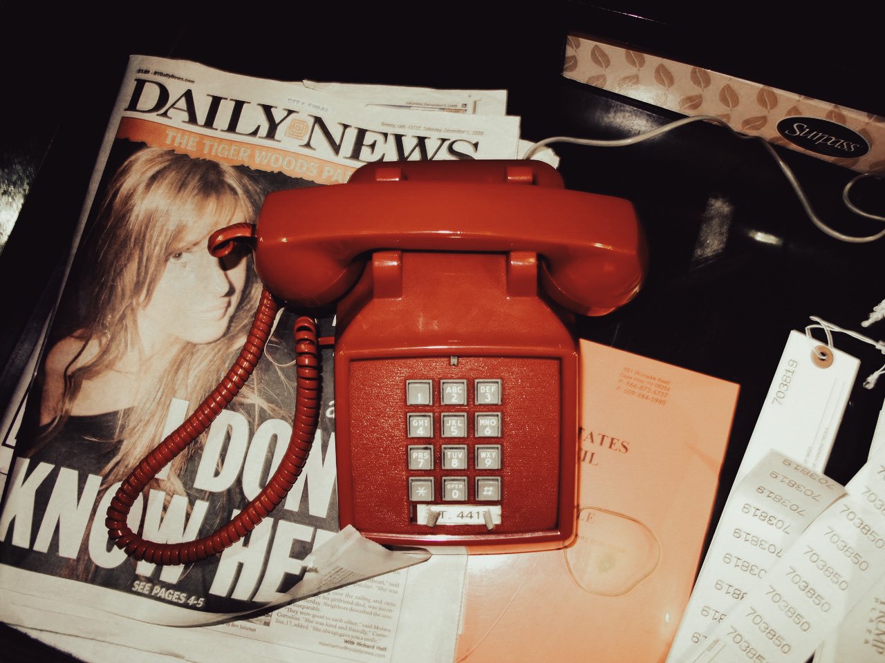 A red rotary phone resting on top of newspapers and receipts on a black surface.