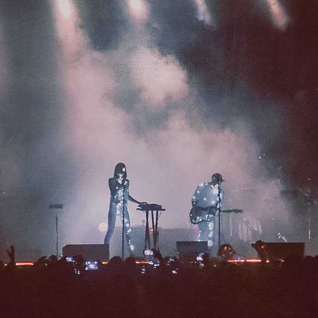Two performers on stage at a concert, with one standing and the other sitting and playing guitar, surrounded by smoke and stage lights, with the audience taking photos.