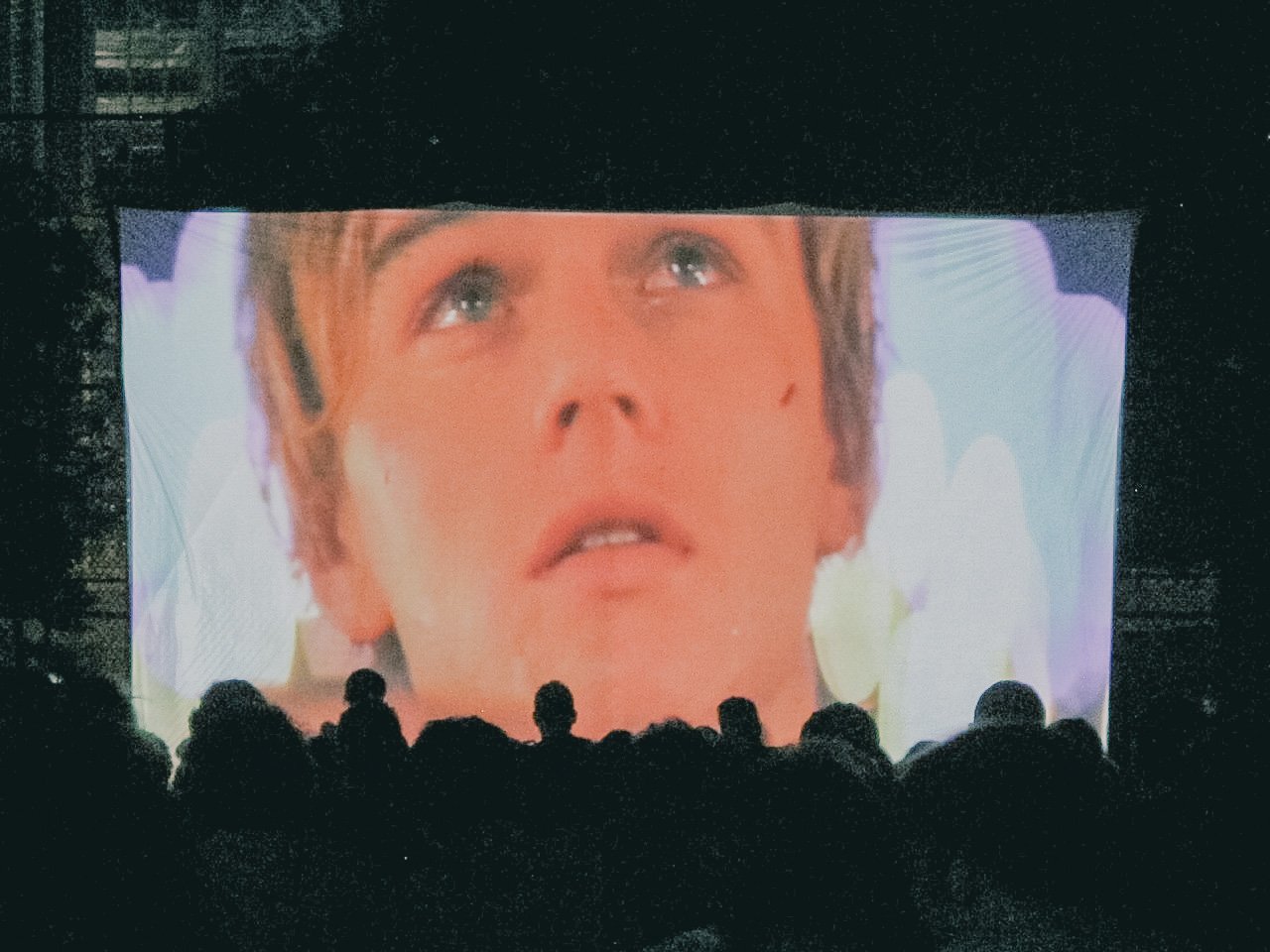 Audience watching a movie or presentation on a large screen showing a close-up of a person's face.