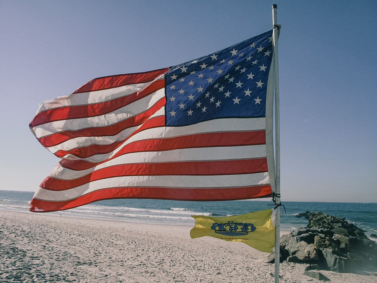 American flag and a yellow flag with a blue emblem on a beach with rocks and ocean in the background.