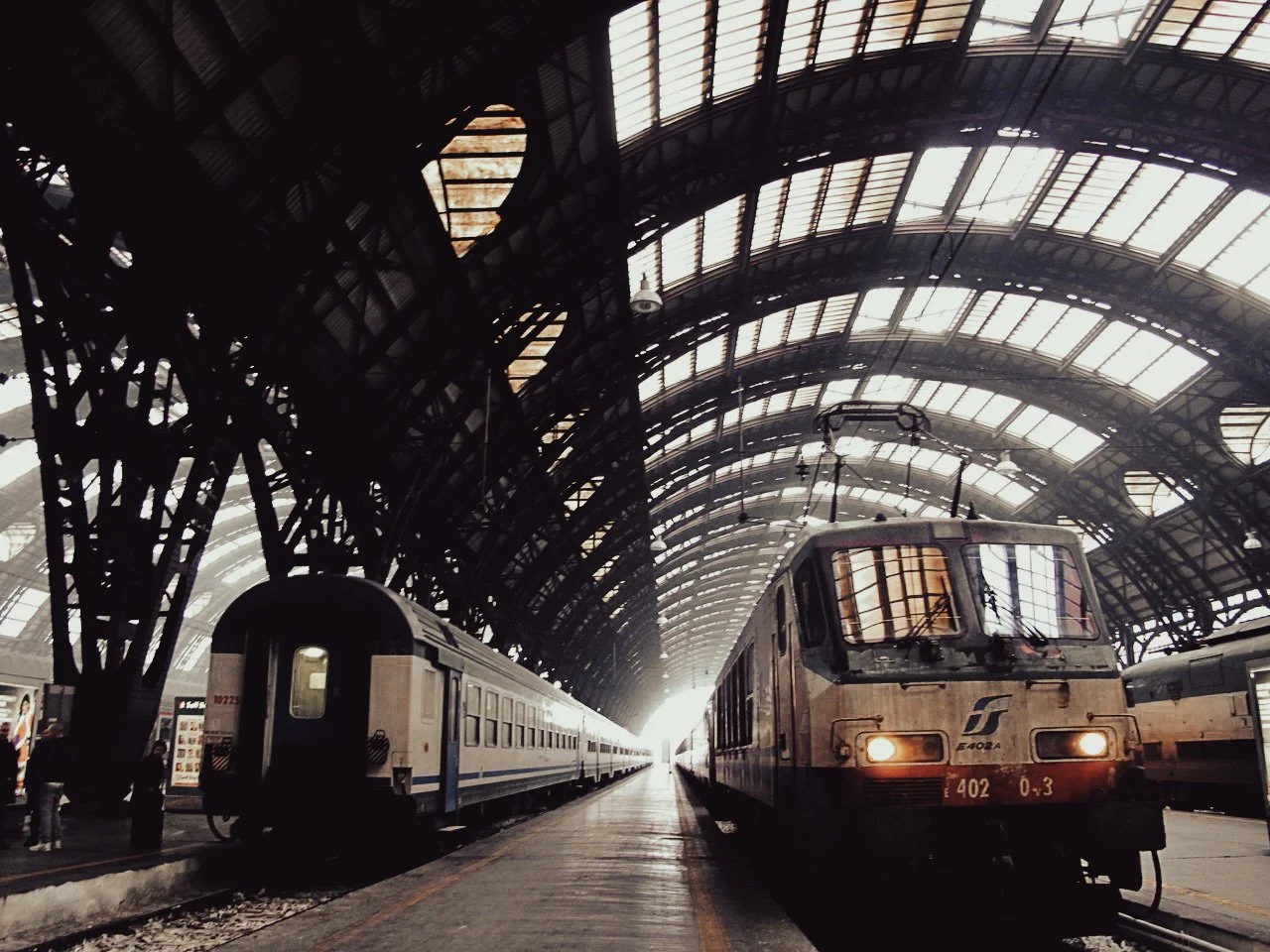 Interior of a train station with two trains, one electric and one passenger, under a large arched glass and metal roof.