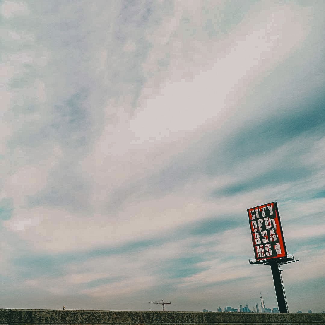 Large digital billboard displaying the words "HEY GOODBYE" in red and white letters, set against a cloudy sky over a city skyline with cranes in the distance.