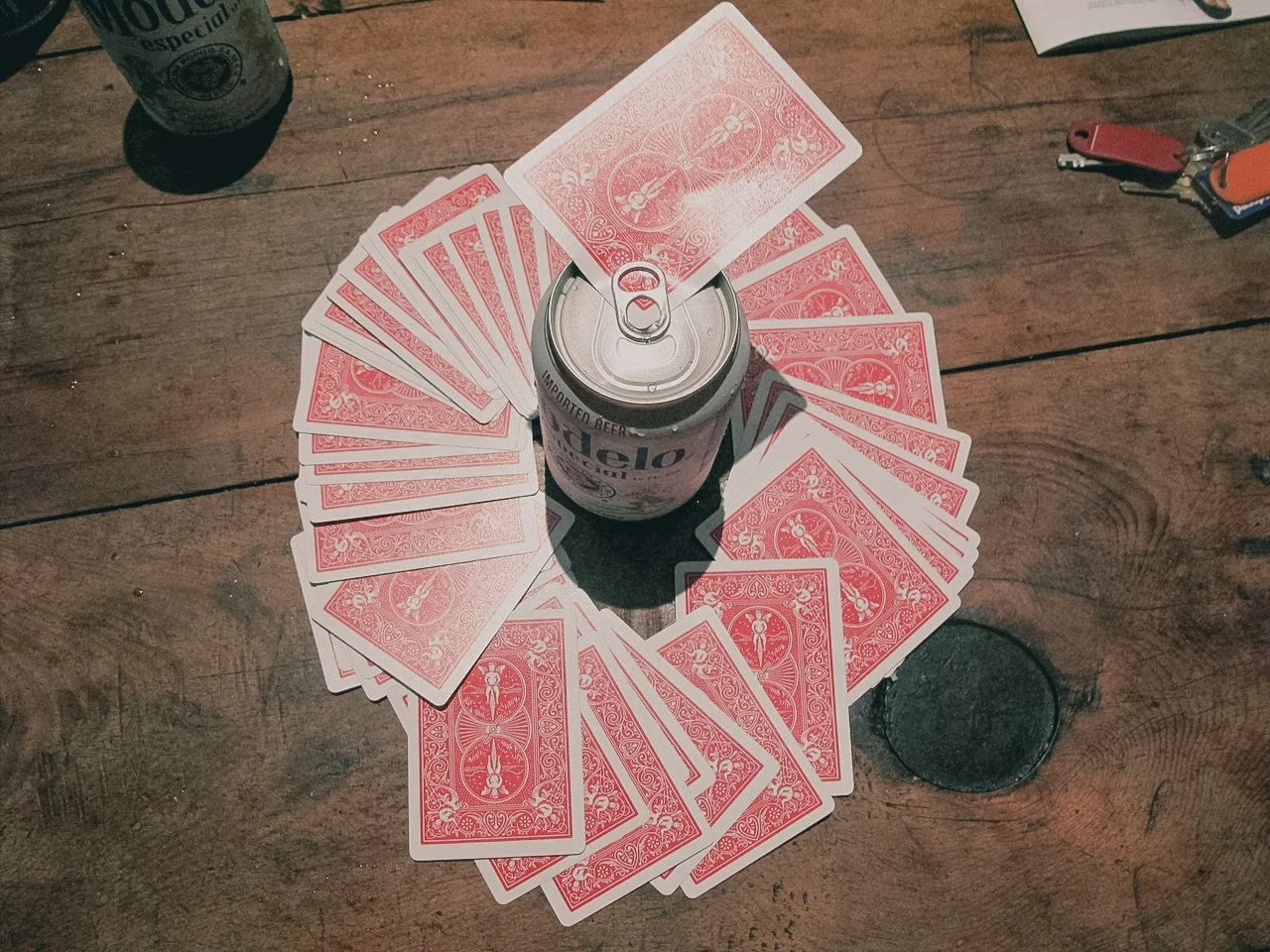 A can of Heilei beer surrounded by a circle of playing cards on a wooden table, with a drink in the background.