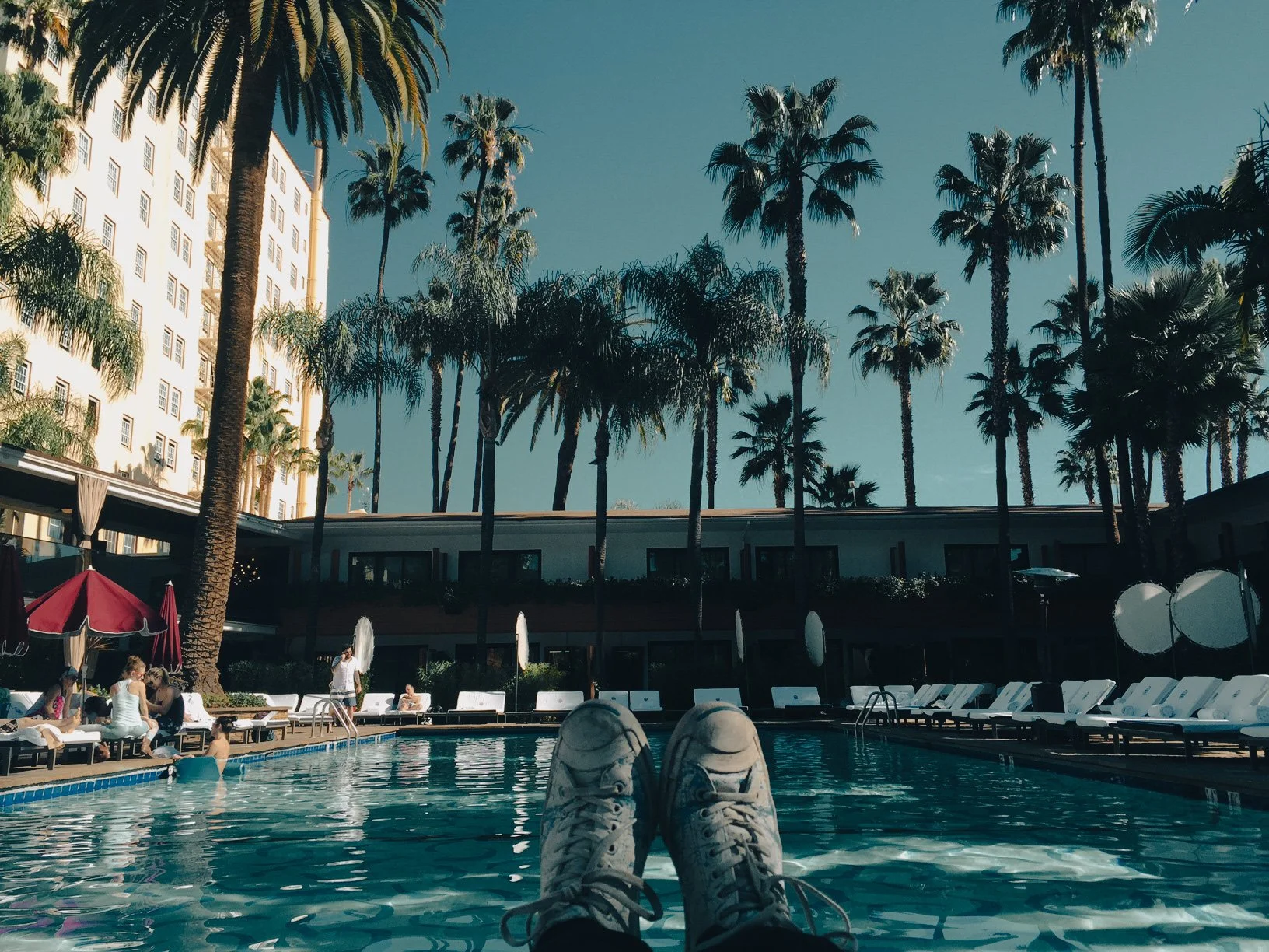 Person relaxing by a pool with white lounge chairs, surrounded by palm trees and a hotel building in the background.